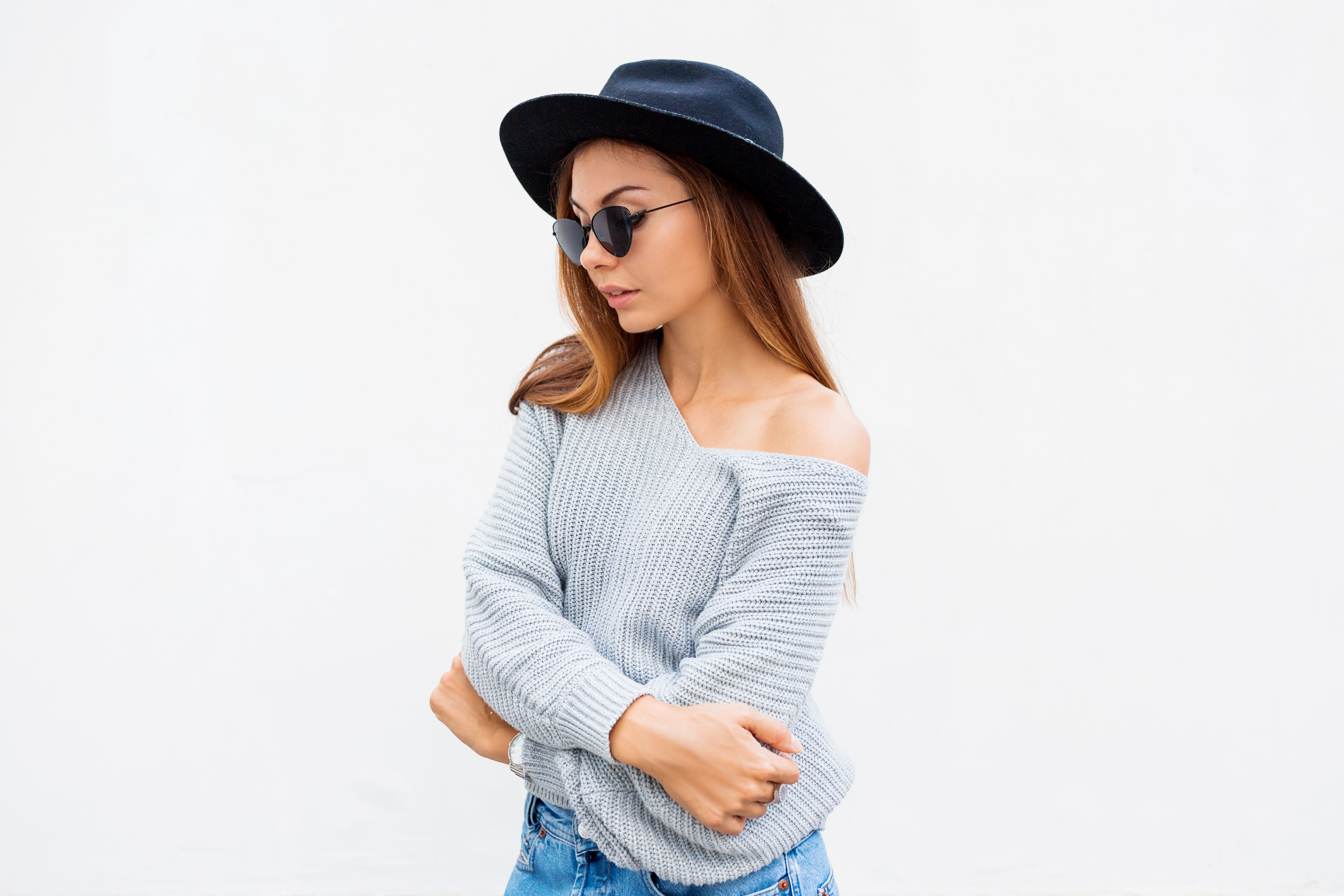 Fashionable portrait of smiling elegant brunette girl in sunglasses , black trendy hat and grey sweater posing over white wall. Bum bag. Positive mood.